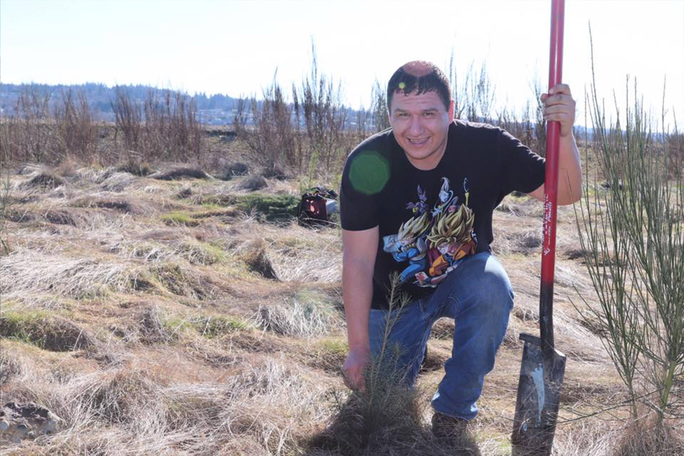 A smiling man kneels next to an evergreen sapling just planted, holding his shovel beside him