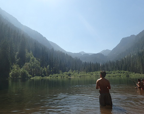 Jacynta Myles Gilford standing in a body of water, fishing with other young people, surrounded by mountains 