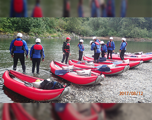 Several canoe-shaped rafts line the shore as a large group of people prepare to traverse the river