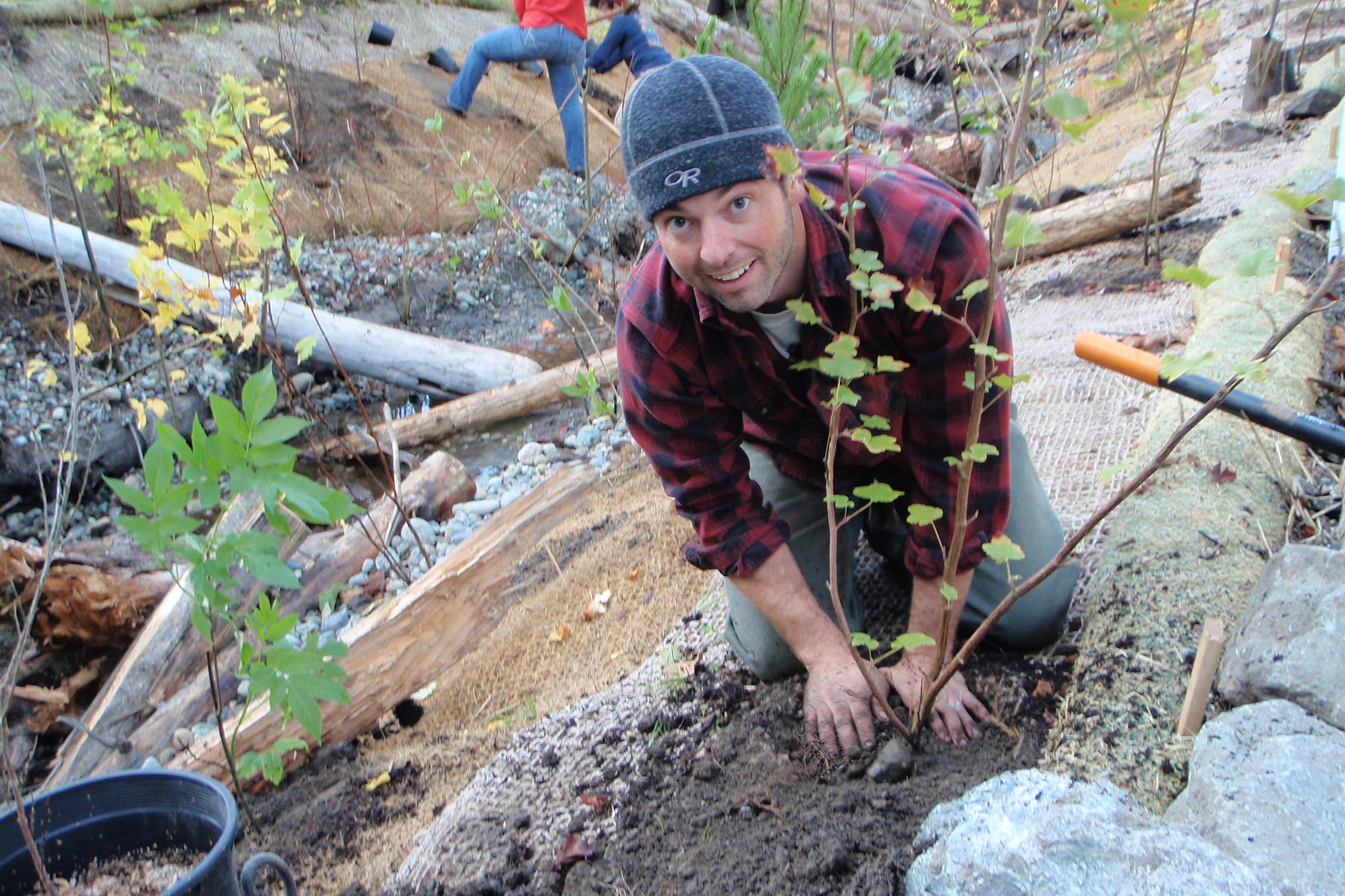 A man smiles as he places dirt at the base of a freshly planted maple tree